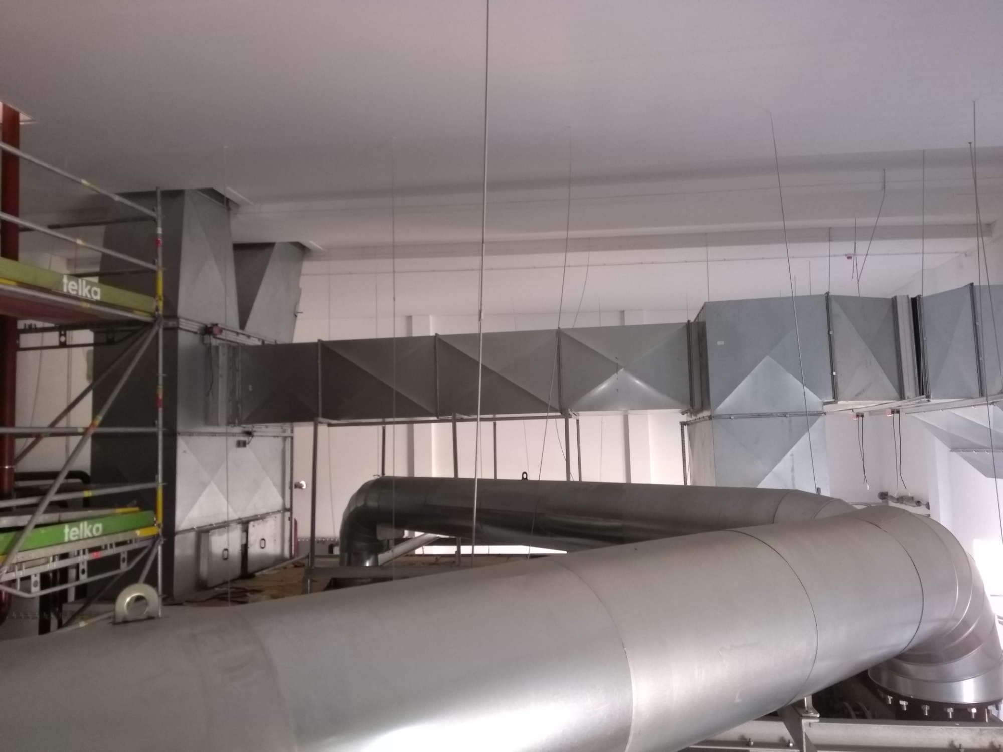 Widok na sufit z zainstalowanymi, srebrnymi przewodami wentylacyjnymi o przekroju okrągłym i kwadratowym, podwieszonymi na cienkich linkach, z widocznym rusztowaniem z boku.