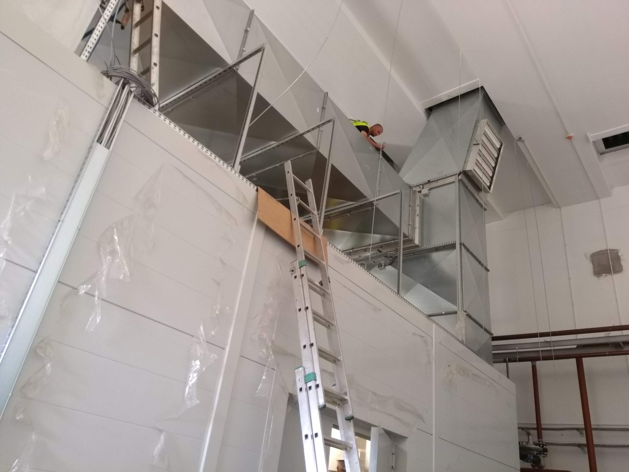 Pracownik w kamizelce odblaskowej montuje system kanałów wentylacyjnych pod sufitem, widoczna drabina i elementy konstrukcyjne.