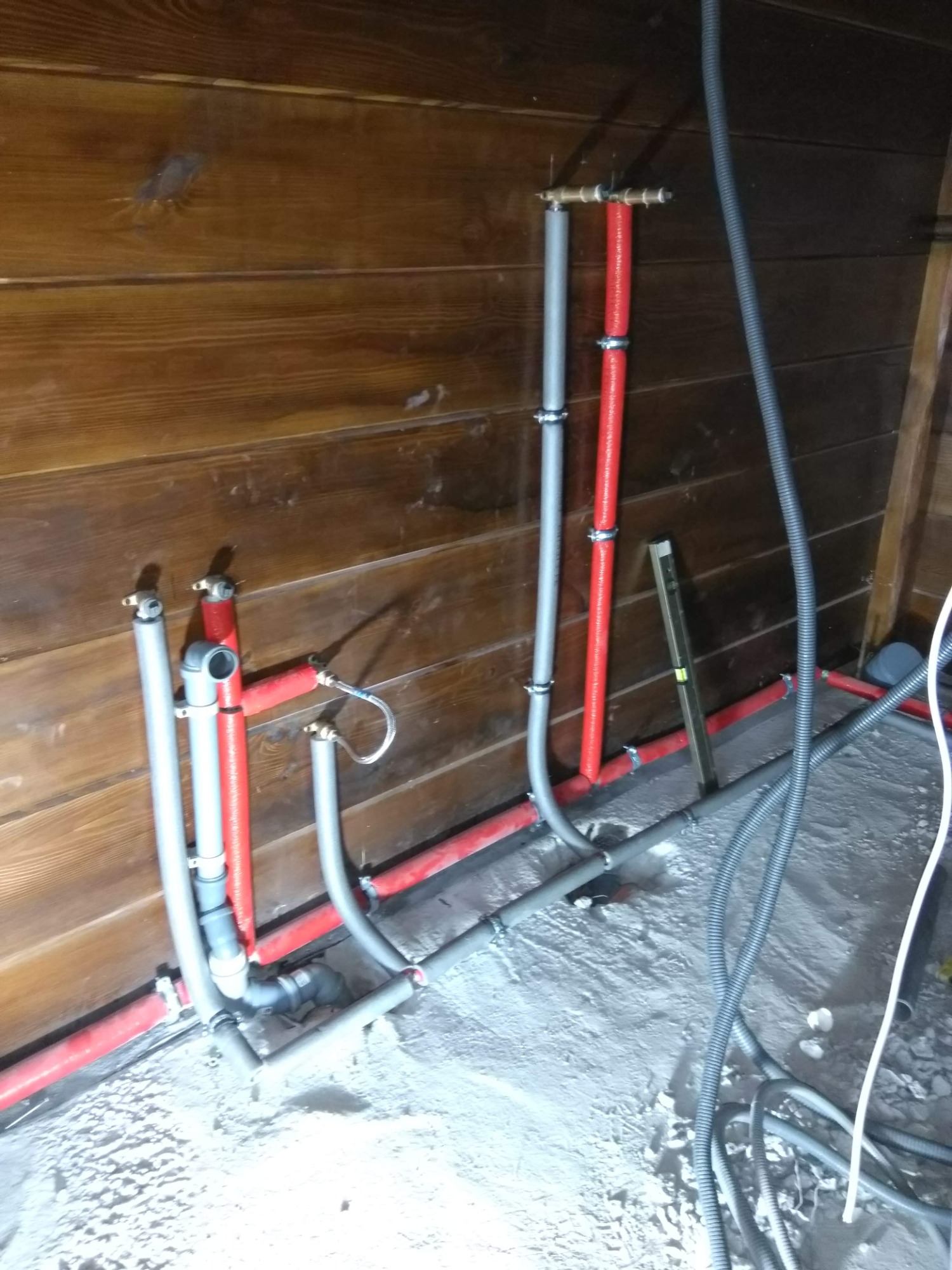 Fragment nowej instalacji wodnej i grzewczej z rur w kolorze czerwonym i szarym, prowadzonej wzdłuż drewnianej ściany w trakcie prac wykończeniowych.