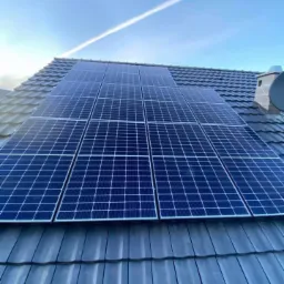 Panele słoneczne zamontowane na ciemnoszarym dachu z dachówki ceramicznej, błękitne niebo z smugą kondensacyjną samolotu w tle.