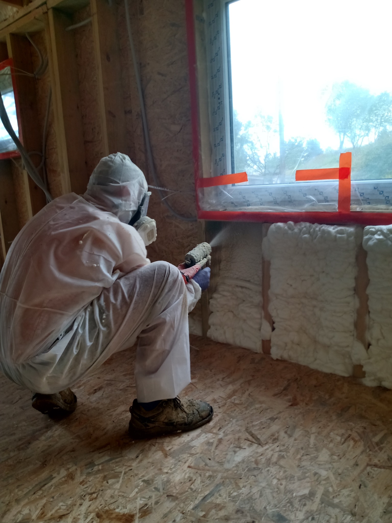Pracownik w kombinezonie ochronnym aplikuje piankę PUR na ścianę wewnątrz budynku, widoczne drewniane elementy konstrukcyjne i okno zabezpieczone taśmą.