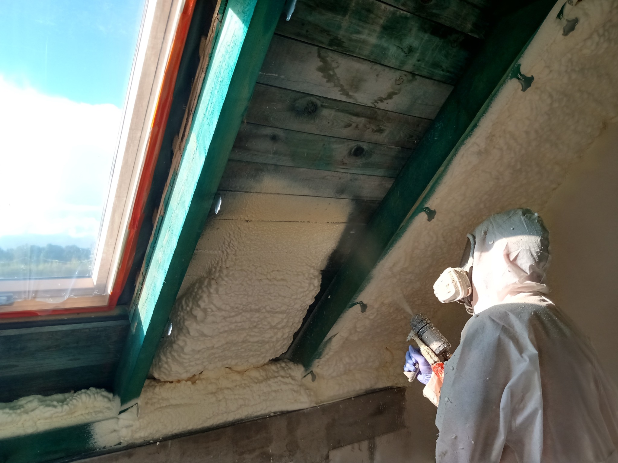 Pracownik w kombinezonie ochronnym aplikuje izolację natryskową na poddaszu, widoczne drewniane elementy konstrukcyjne i okno dachowe z widokiem na błękitne niebo.