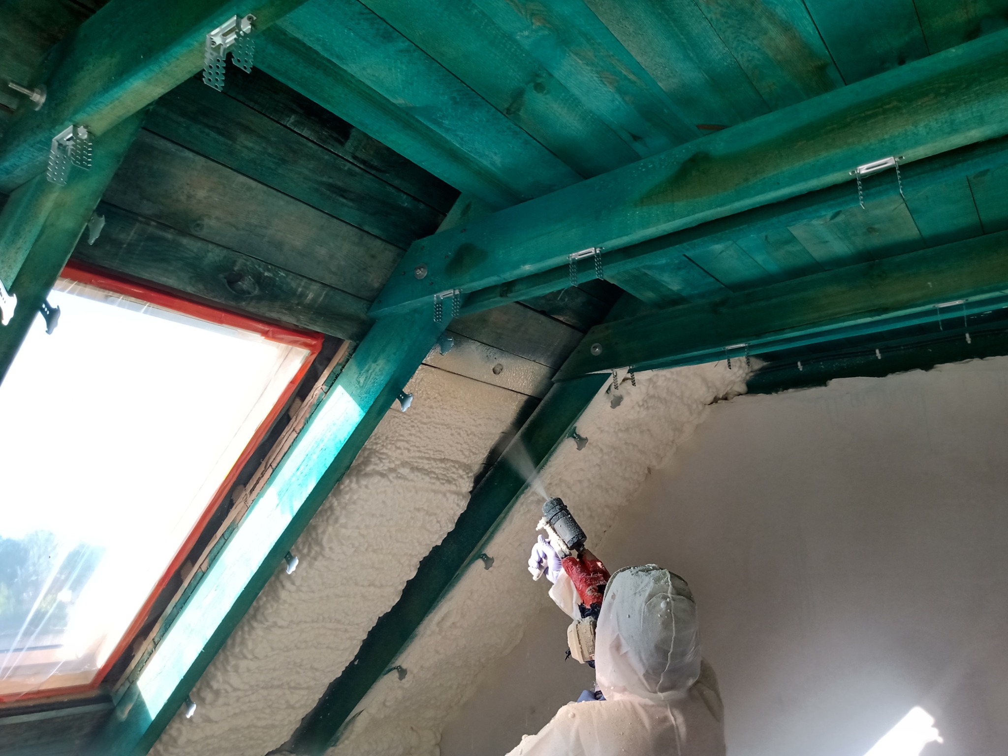 Pracownik w kombinezonie ochronnym natryskuje białą pianę izolacyjną na poddasze z drewnianą konstrukcją pomalowaną na zielono, widoczne okno dachowe.