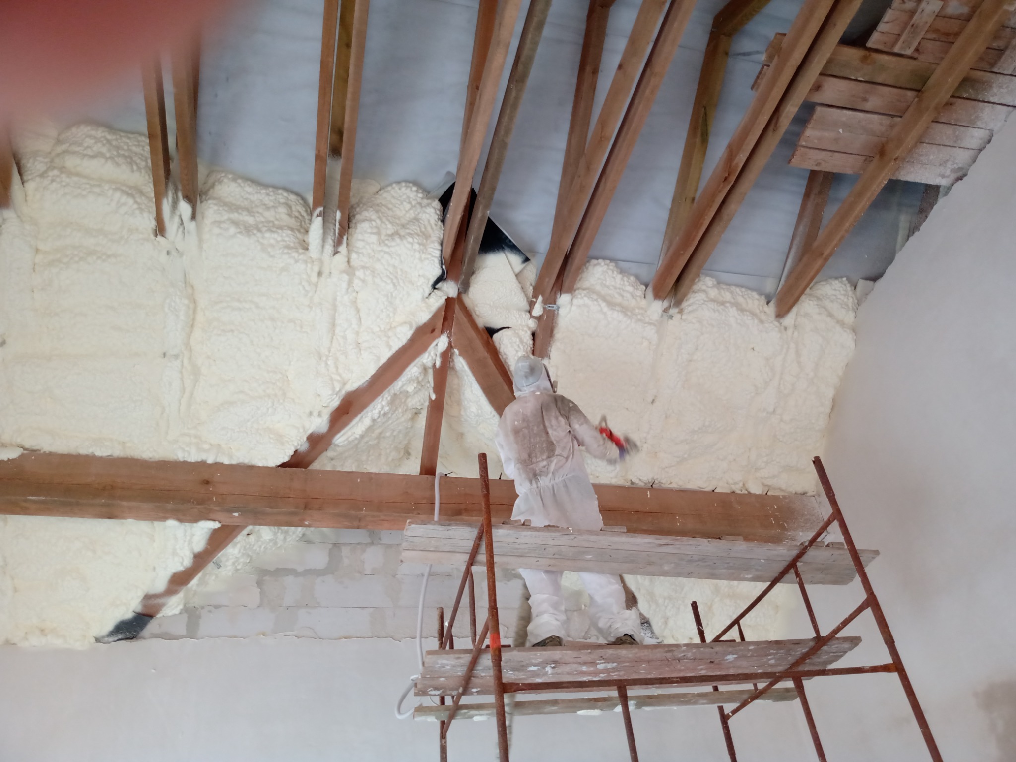 Pracownik w kombinezonie ochronnym aplikuje piankę izolacyjną na poddaszu, widoczne drewniane krokwie i rusztowanie.