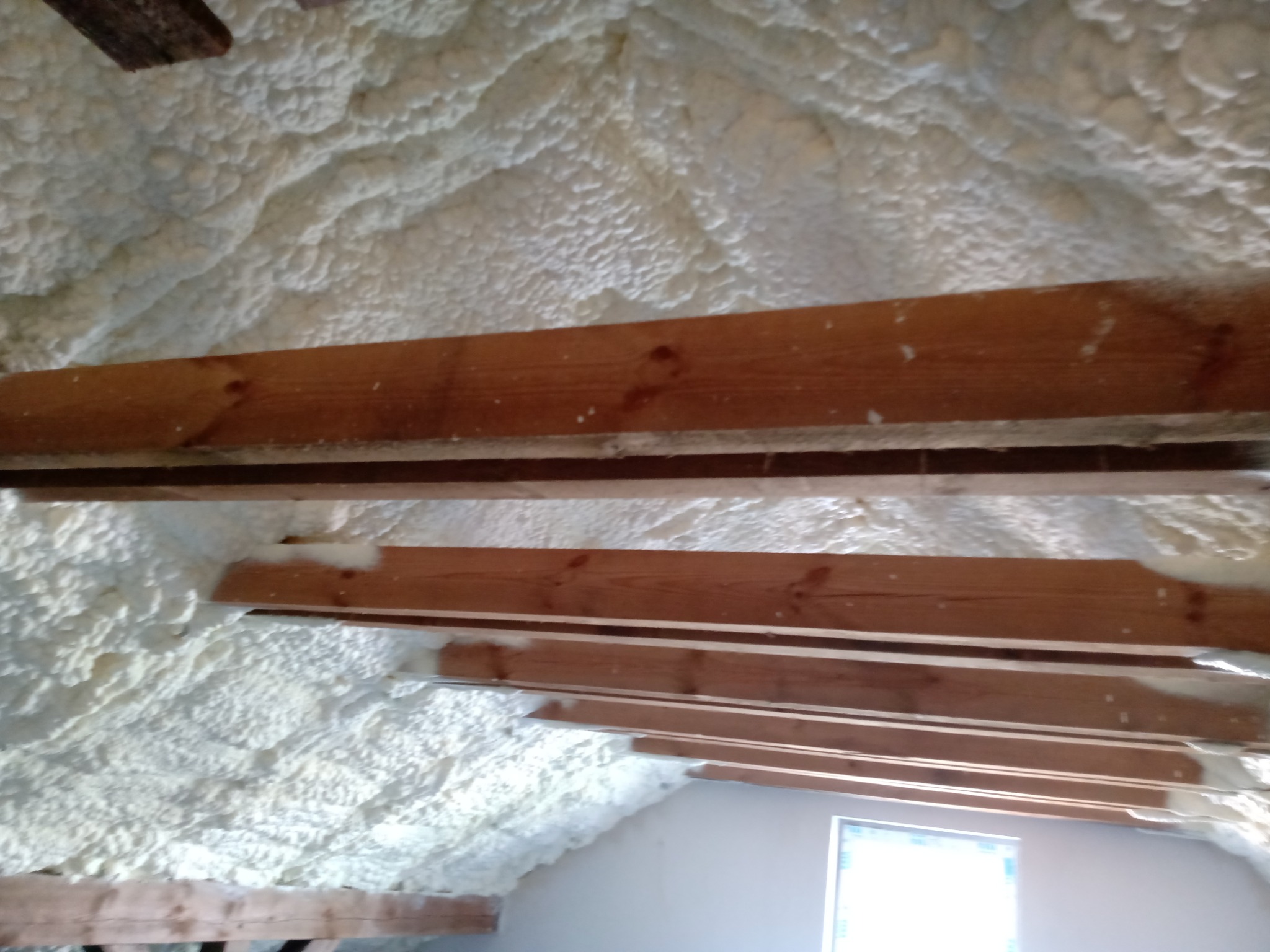 Poddasze ocieplone białą pianką, widoczne drewniane belki konstrukcyjne, okno w tle.