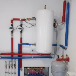 Instalacja centralnego ogrzewania z białym zasobnikiem ciepłej wody, rozdzielaczem podłogowym i rurami w izolacji czerwonej i niebieskiej w nowym pomieszczeniu