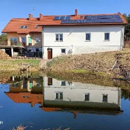 Dwukondygnacyjny dom z panelami słonecznymi na dachu, odbijający się w stawie, obok sterta drewna