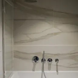 Wykończona łazienka z marmurowymi płytkami na ścianach, chromowaną armaturą i szklanym panelem prysznicowym przy wannie.