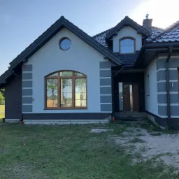 Nowoczesny dom jednorodzinny z szarą elewacją, drewnianą stolarką okienną i ciemną dachówką, widok z zewnątrz w słoneczny dzień, teren wokół domu jeszcze niezagospodarowany.