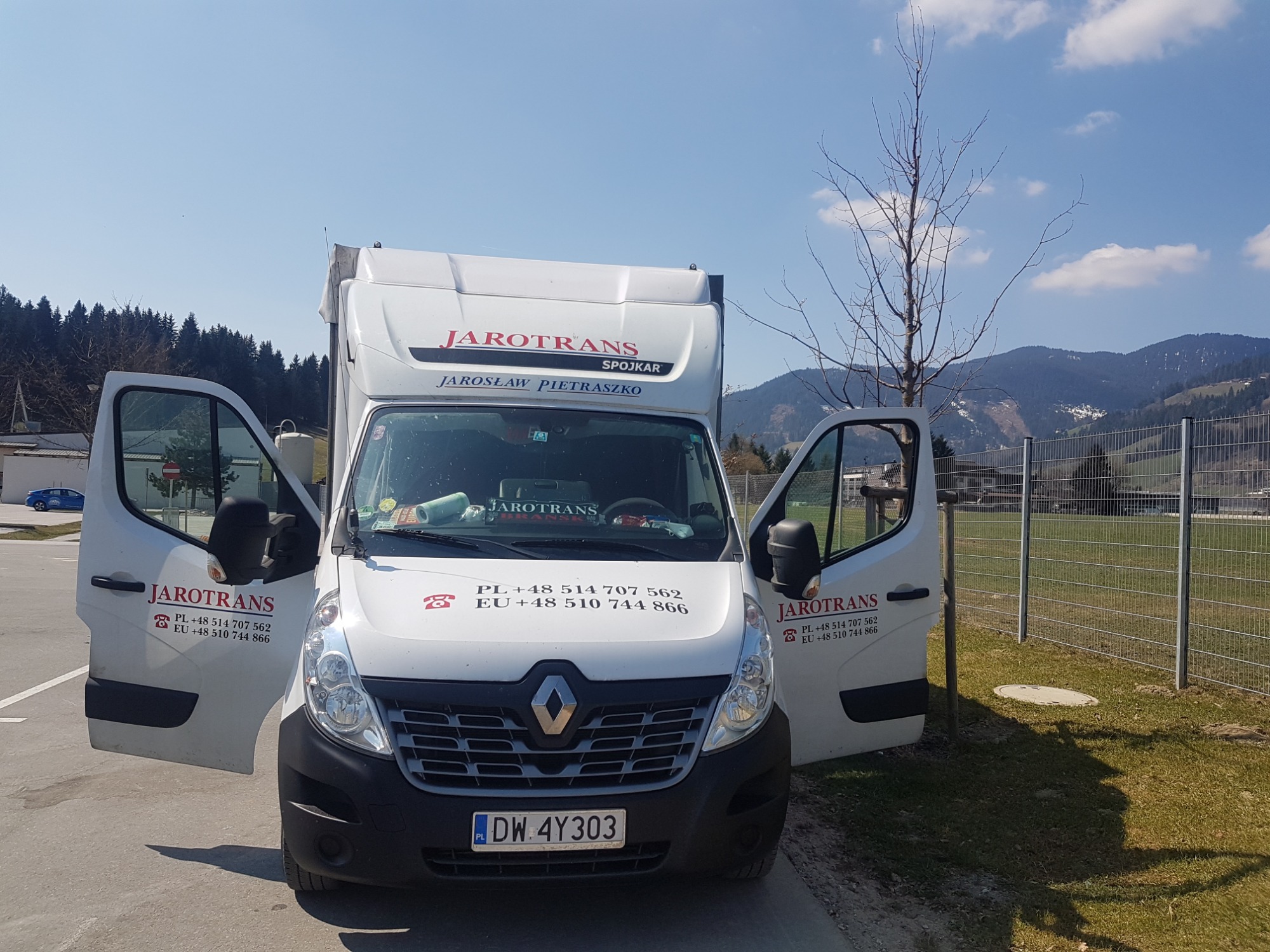 Biały bus firmy Jarotrans z otwartymi drzwiami na parkingu z widokiem na góry w tle.