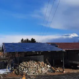 Instalacja paneli słonecznych na zadaszeniu w wiejskim otoczeniu, obok sterta kamieni i częściowo ośnieżony teren.
