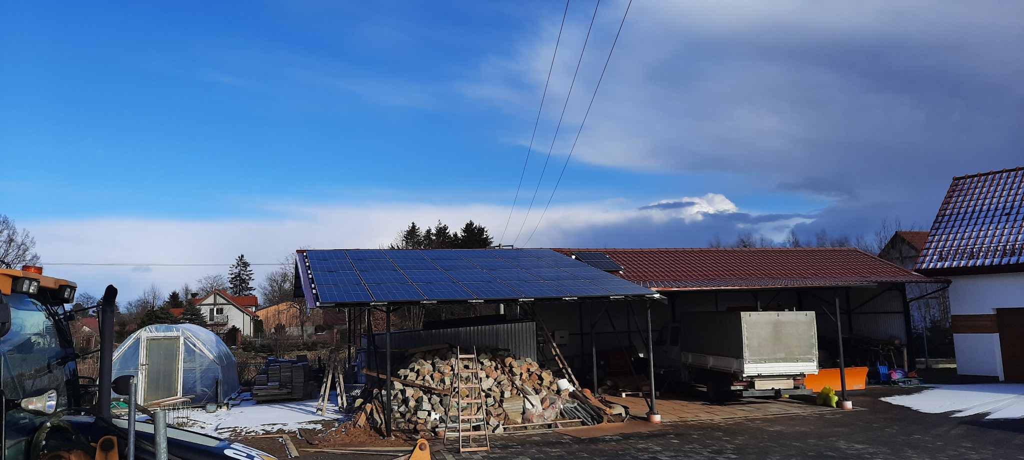 Instalacja paneli słonecznych na zadaszeniu w wiejskim otoczeniu, obok sterta kamieni i częściowo ośnieżony teren.