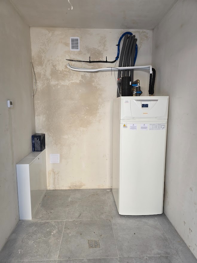 Biała pompa ciepła w surowym pomieszczeniu z widocznymi rurami instalacji, wentylacją i szarą podłogą z płytek. Ściany w stanie deweloperskim.