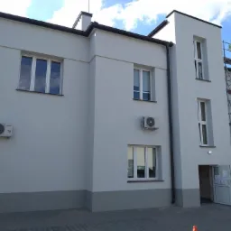 Budynek biurowy po remoncie elewacji, widoczne okna z białymi ramami, klimatyzatory na ścianie, rusztowanie przy wejściu i szara podmurówka.