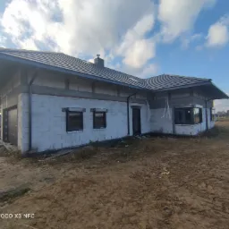 Dom murowany w trakcie budowy, widoczne białe bloczki, czarna dachówka, okna z roletami, teren wokół w stanie surowym.