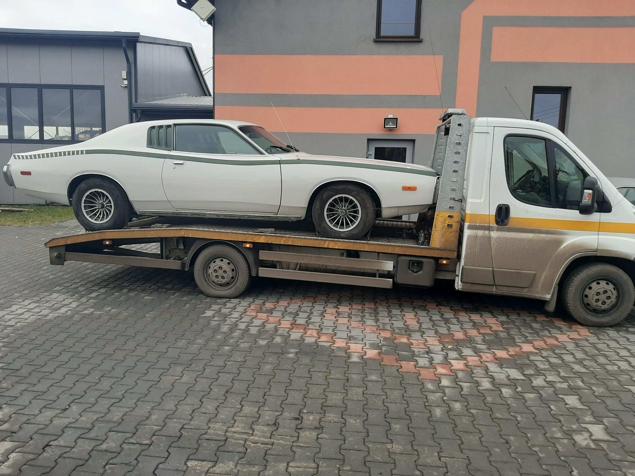 Biały klasyczny samochód z zielonym pasem transportowany na platformie pojazdu pomocy drogowej, widoczny na tle budynku z elementami w kolorze brzoskwiniowym.