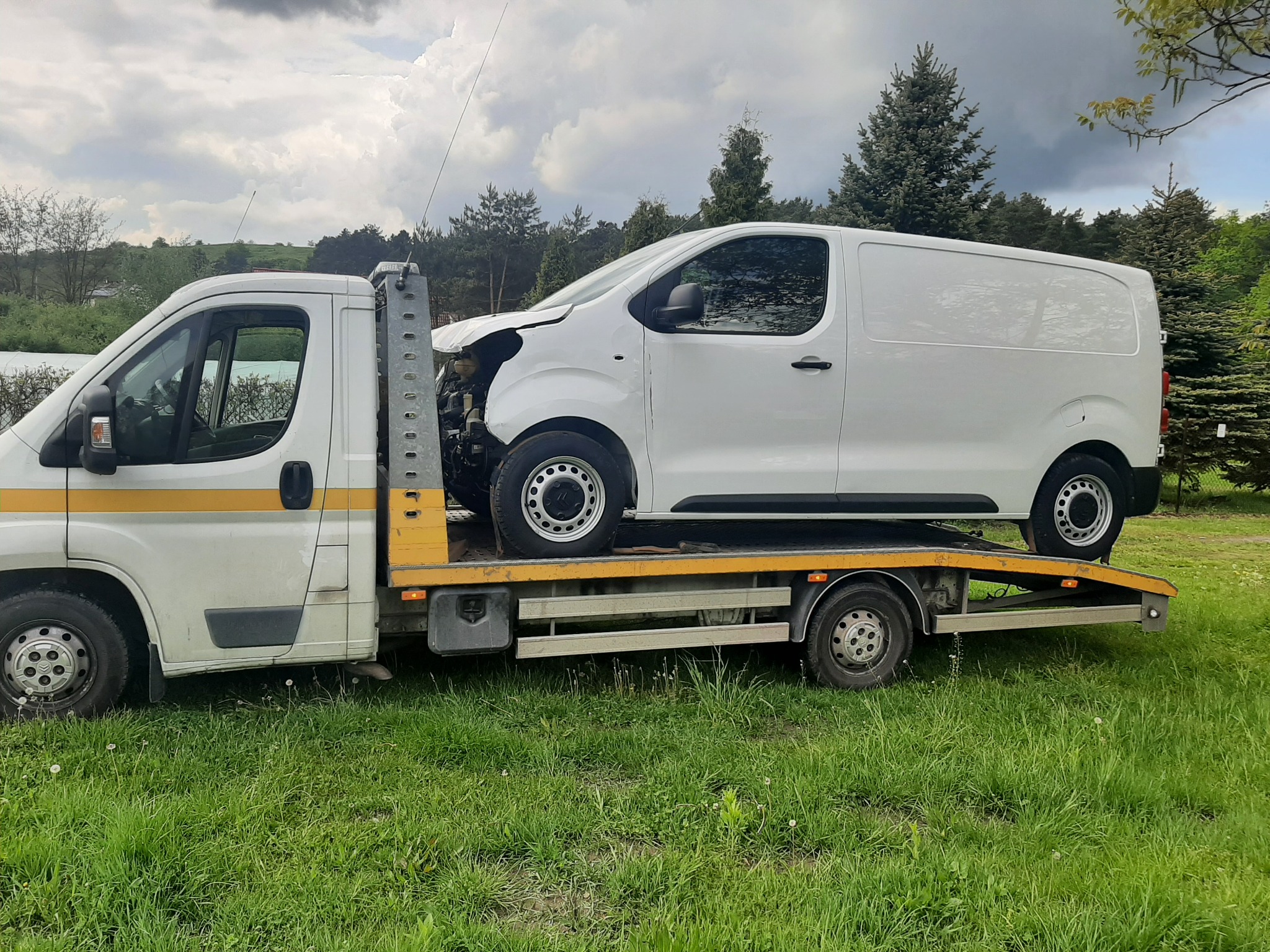 Uszkodzony biały samochód dostawczy na żółtej platformie holowniczej, widok z boku na tle zielonej trawy i pochmurnego nieba.