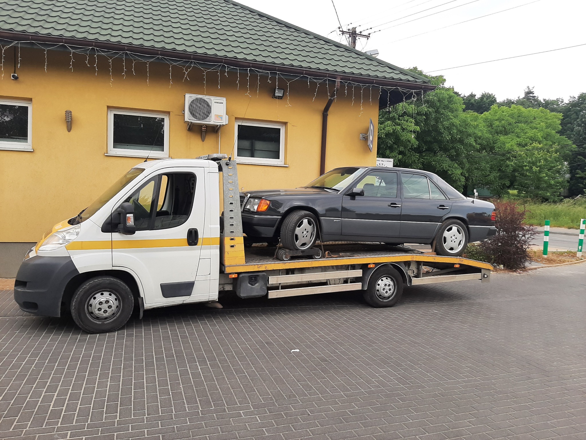 Laweta z załadowanym ciemnoszarym Mercedesem W124 na tle żółtego budynku z klimatyzacją i zielonym dachem.