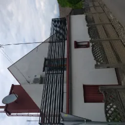 Biały budynek z czarną, metalową balustradą balkonu, ozdobnymi, ażurowymi elementami przy chodniku, anteną satelitarną i klimatyzatorem na elewacji.