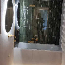 Nowoczesna łazienka z zielonymi, pionowymi płytkami, złotą armaturą, podświetlanym lustrem i podświetleniem pod wanną, z widocznym odbiciem osoby w szklanej ściance wanny.