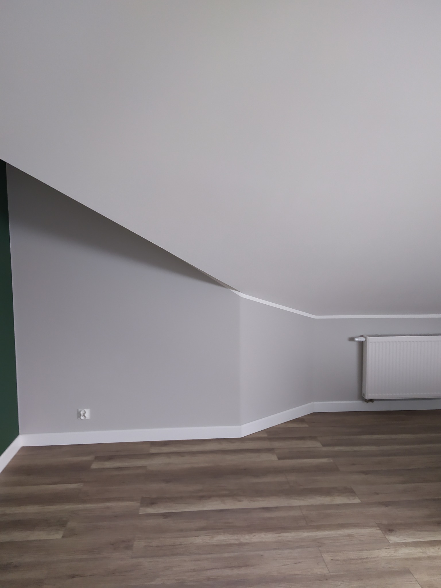 Poddasze po remoncie: szare ściany, biały sufit i listwy przypodłogowe, drewnopodobna podłoga, grzejnik na ścianie, gniazdko elektryczne.