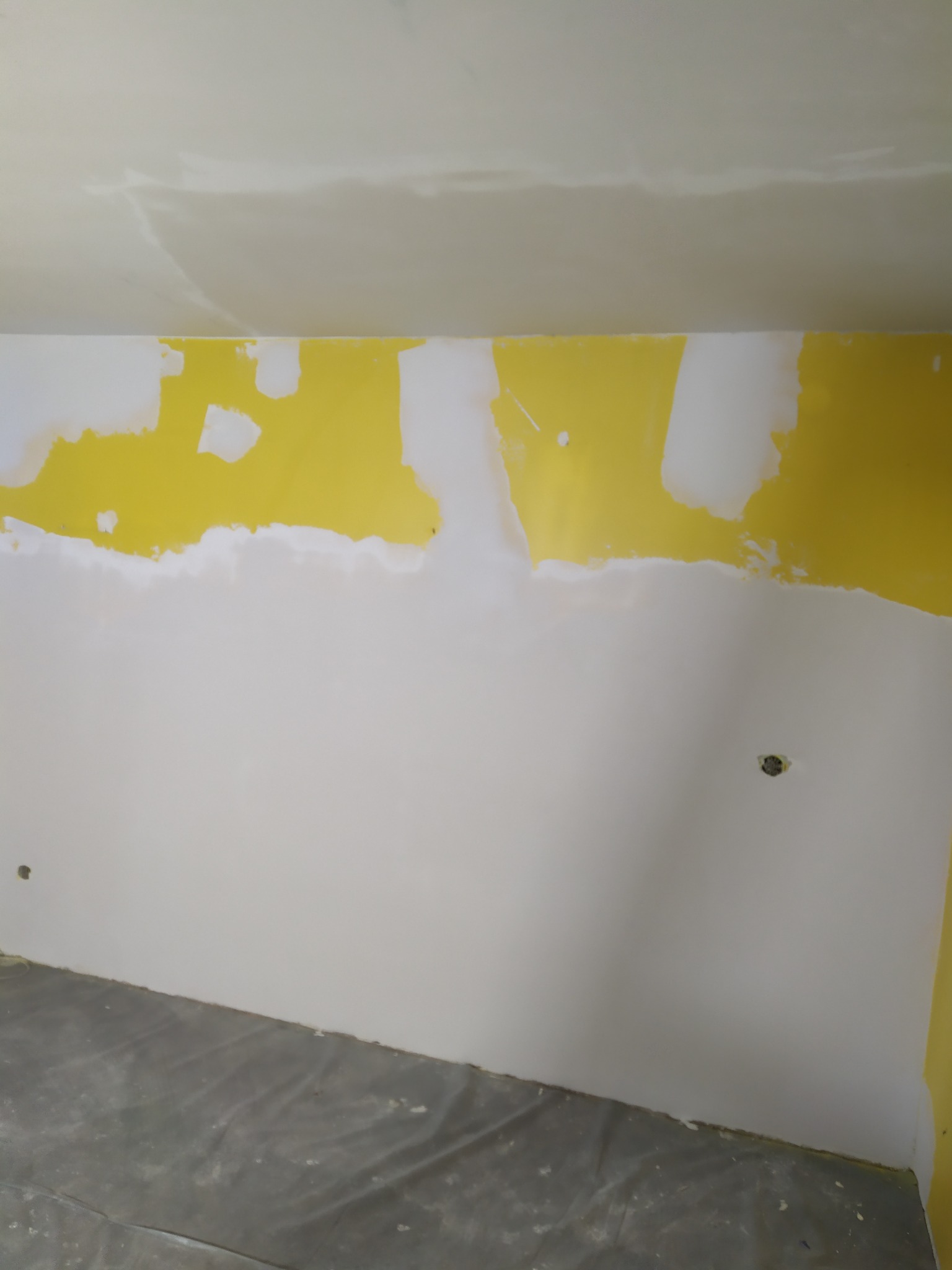 Ściana w trakcie szpachlowania, widoczne fragmenty żółtej farby pod białą szpachlą, zabezpieczona podłoga folią.