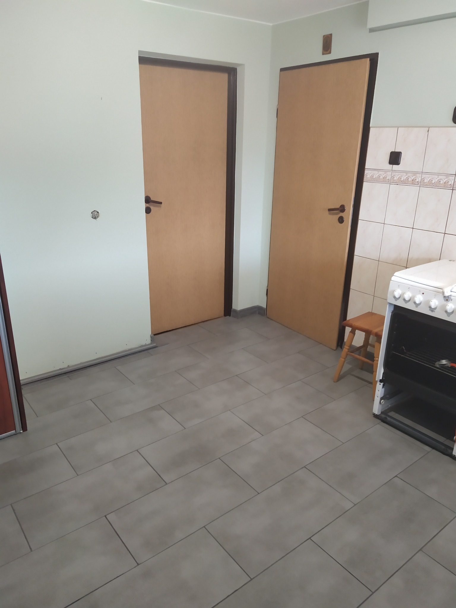 Kuchnia po remoncie: szare płytki podłogowe, dwie drewniane drzwi, biała kuchenka z taboretem obok. Widoczne ślady po gniazdku na ścianie.