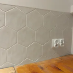Ściana wyłożona heksagonalnymi, jasnymi płytkami ceramicznymi z widocznym gniazdkiem elektrycznym i fragmentem drewnianego blatu.