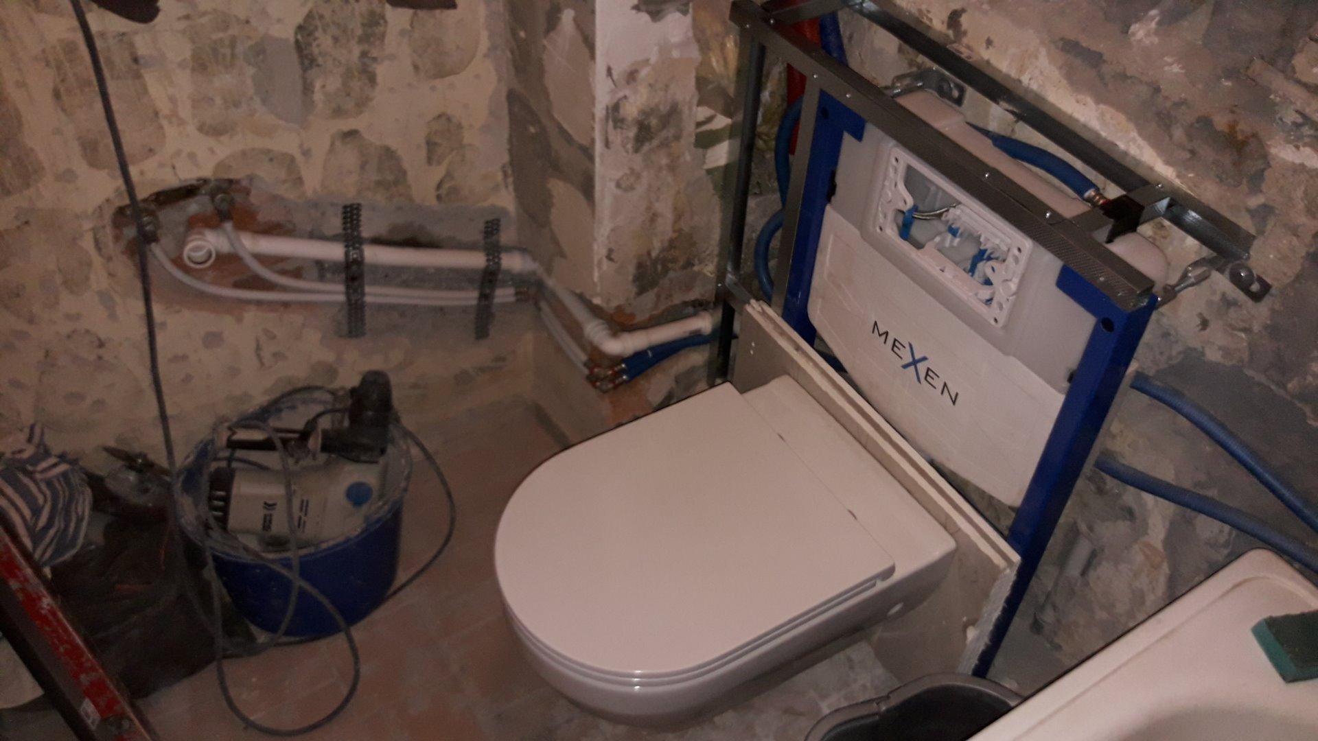 Montaż podtynkowego stelaża WC marki Mexen w surowej łazience z kamiennymi ścianami, widoczne rury instalacyjne i narzędzia w wiadrze.