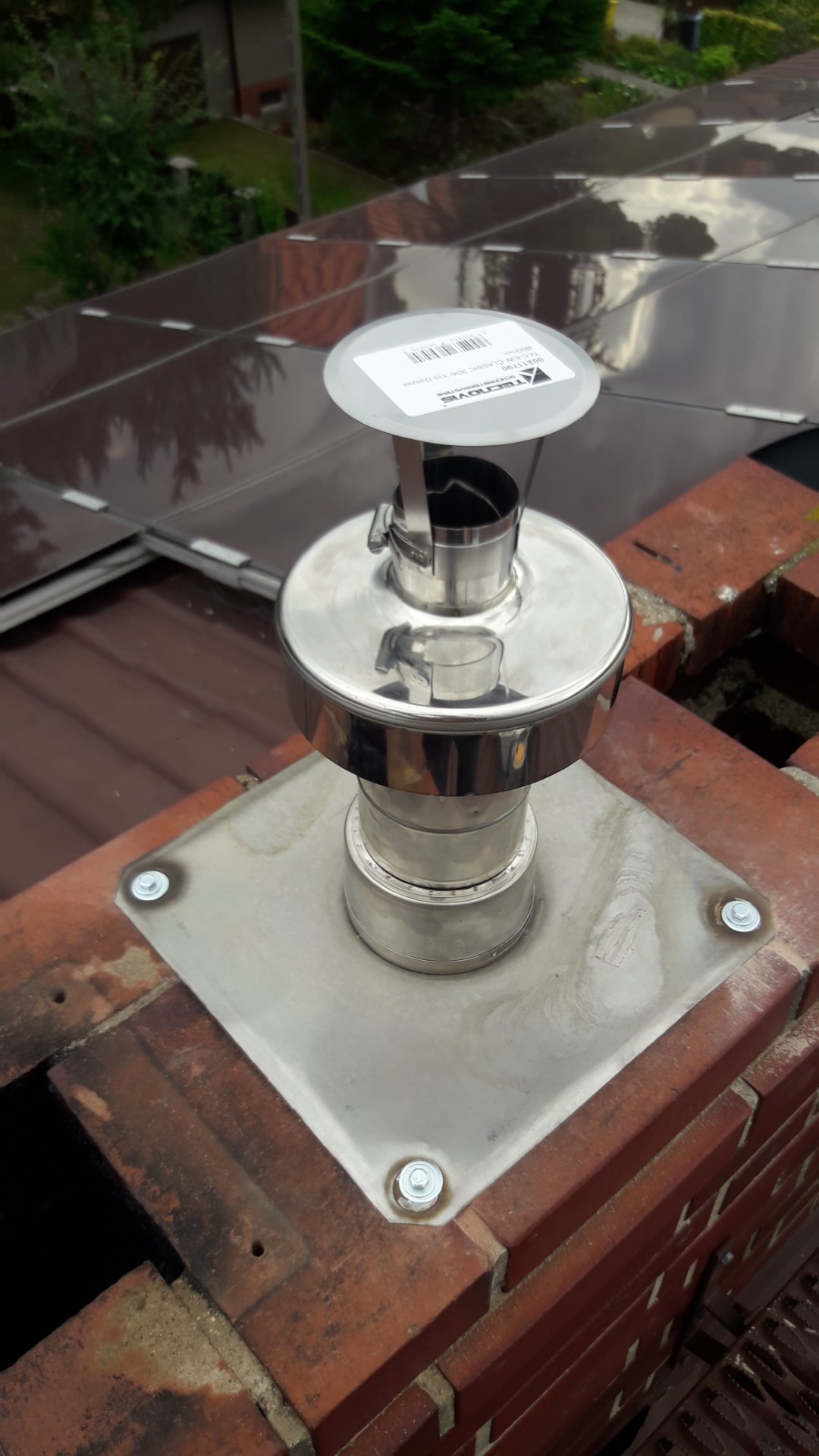 Nowy, błyszczący, metalowy komin wentylacyjny zamontowany na ceglanym kominie, widoczne panele słoneczne w tle.