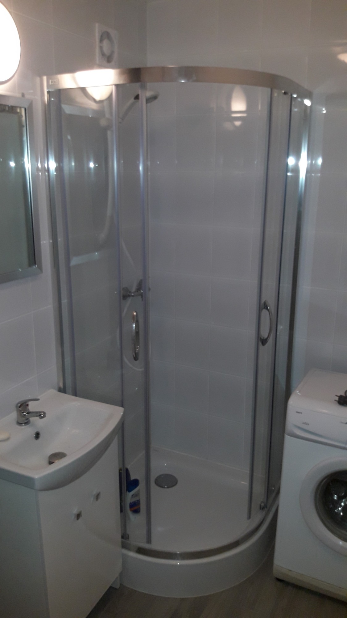 Biała kabina prysznicowa narożna z chromowanymi profilami, umywalka z szafką i pralka w małej łazience z jasnymi płytkami.