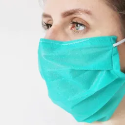 Portret kobiety z profilu, noszącej turkusową, plisowaną maseczkę ochronną na twarzy, z widocznym białym szwem i elastycznymi gumkami za uszami, na białym tle.