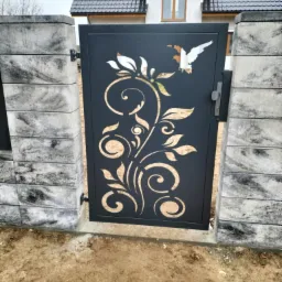 Czarna, ażurowa furtka z motywem roślinnym i ptaków, osadzona między betonowymi słupkami. W tle dom z szarą dachówką.