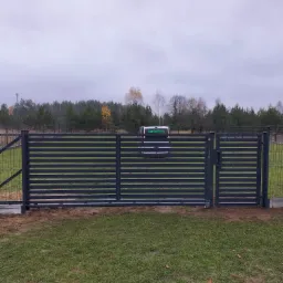 Nowoczesna, pozioma brama wjazdowa z metalowych profili w kolorze grafitowym, częściowo otwarta, z widocznym zamkiem, na tle zielonego trawnika i zalesionego krajobrazu w pochmurny dzień. W tle...