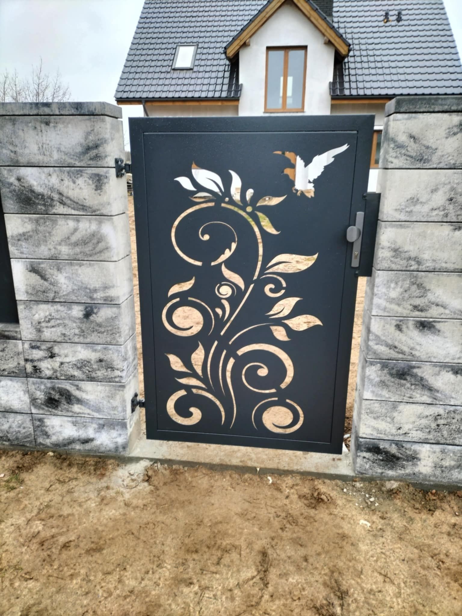 Czarna, ażurowa furtka z motywem roślinnym i ptaków, osadzona między betonowymi słupkami. W tle dom z szarą dachówką.