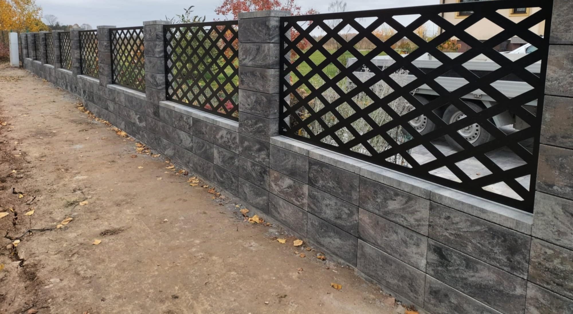 Nowoczesne ogrodzenie z szarego kamienia łupanego i czarnych, ażurowych paneli. Widok z boku, jesienne liście na ziemi. W tle budynek mieszkalny i zaparkowany pojazd.
