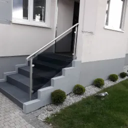 Zewnętrzne schody wejściowe z ciemnymi stopniami i balustradą ze szkła oraz stali nierdzewnej, widok na elewację domu z oknami i ozdobnymi krzewami na tle trawnika z figurką ptaka.