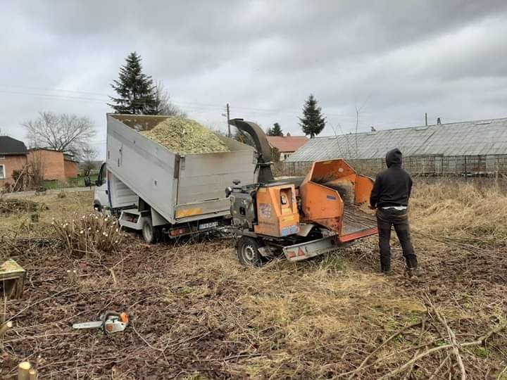 Pomarańczowy rębak do gałęzi przy ciężarówce wypełnionej zrębkami, obok leży piła łańcuchowa, w tle budynki i pochmurne niebo.