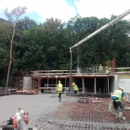 Wylewanie betonu na zbrojoną konstrukcję stropu budynku, widoczni pracownicy w kaskach i kamizelkach odblaskowych, pompa do betonu w tle, otoczenie drzewa.