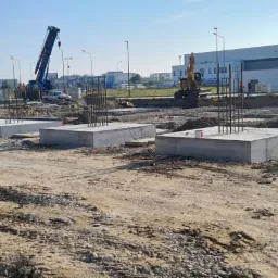 Betonowe stopy fundamentowe z wystającymi prętami zbrojeniowymi na placu budowy, w tle widoczny dźwig i budynek przemysłowy.