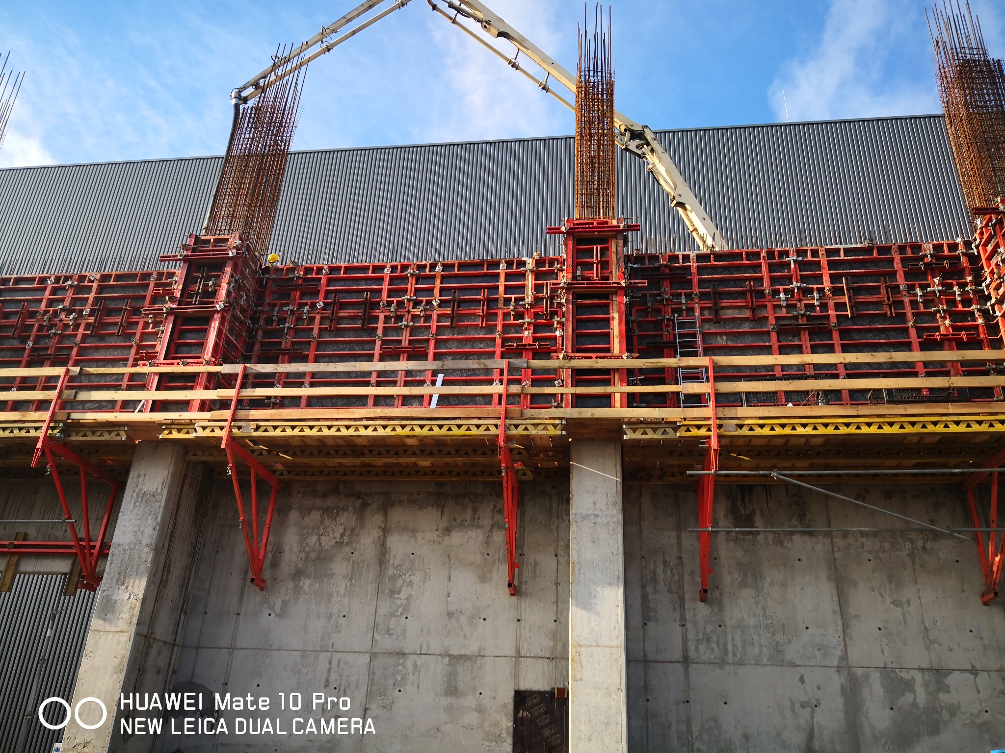Szare, betonowe ściany szalunkowe z wystającymi pionowymi prętami zbrojeniowymi, podtrzymywane przez czerwone, metalowe podpory i drewniane elementy konstrukcyjne, z ramieniem pompy do betonu w tle.