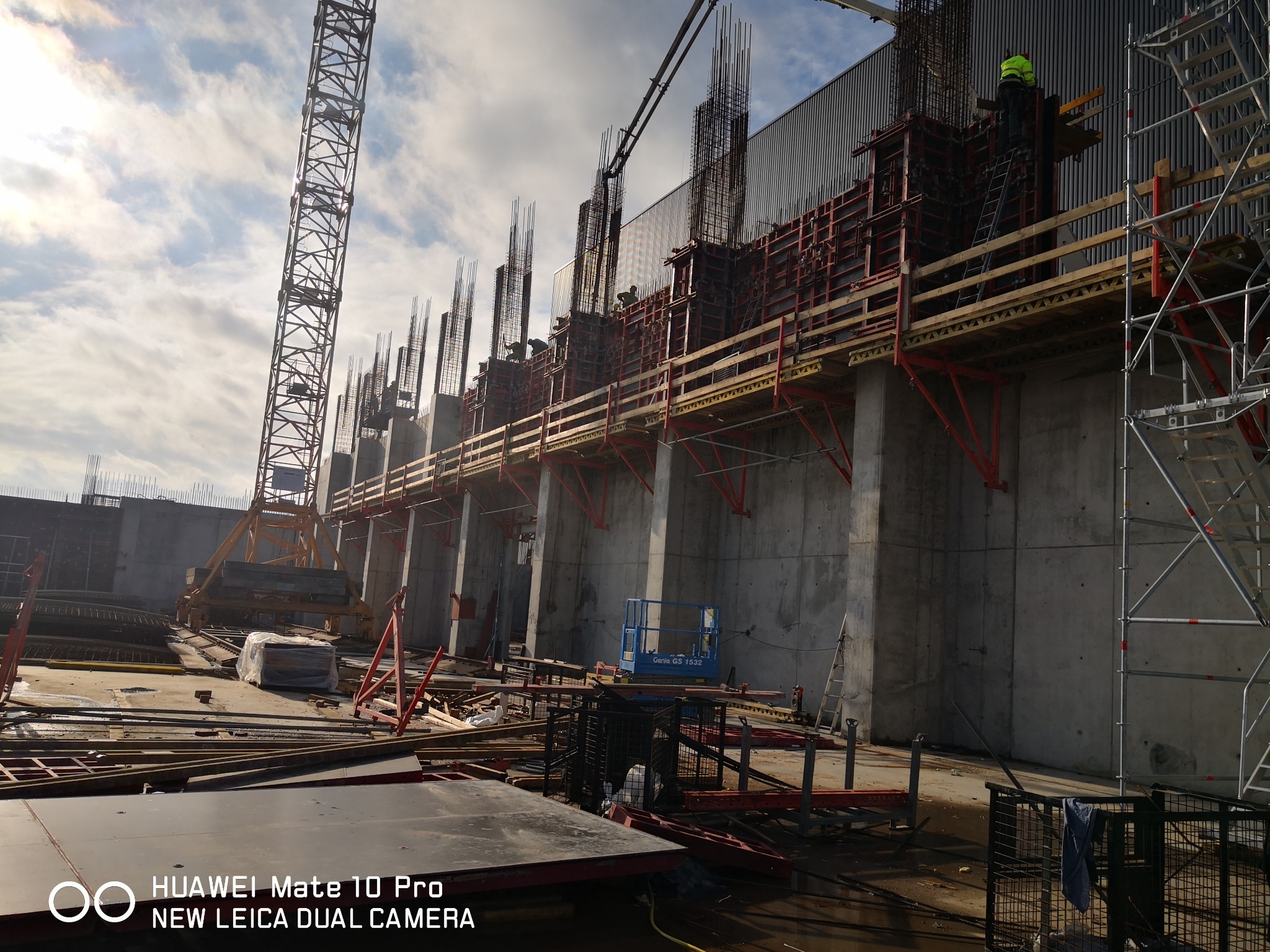 Wysokie ściany szalunkowe z wystającą stalą zbrojeniową na placu budowy, widoczne rusztowania, żuraw i podnośnik koszowy.