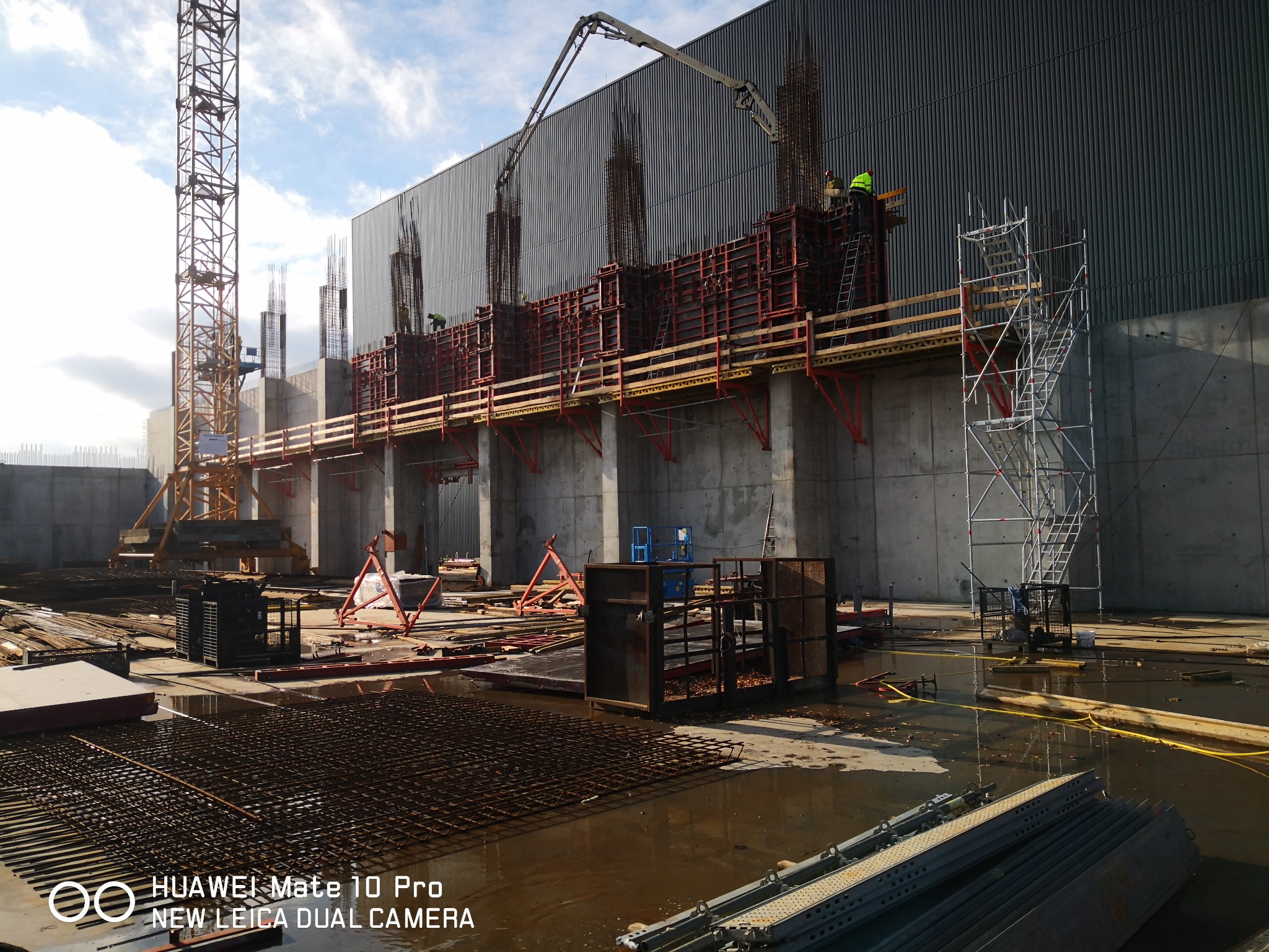 Wysoka konstrukcja w trakcie budowy z betonu i stali, widoczne deskowanie, dźwig i pompa do betonu, teren budowy częściowo zalany wodą, pracownicy w kamizelkach odblaskowych na górze konstrukcji.