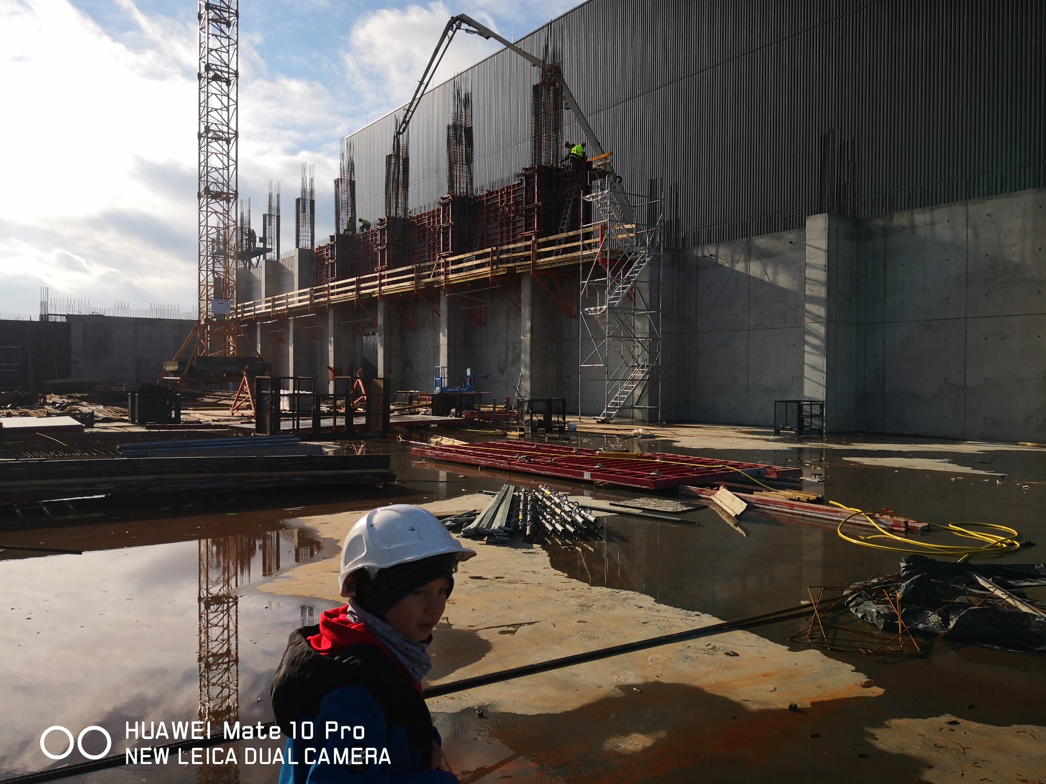 Budowa hali przemysłowej: widok na zbrojenie pionowych słupów i betonowe fundamenty w trakcie wylewania betonu z pompy, w tle żuraw wieżowy, na pierwszym planie pracownik w kasku i kałuże.