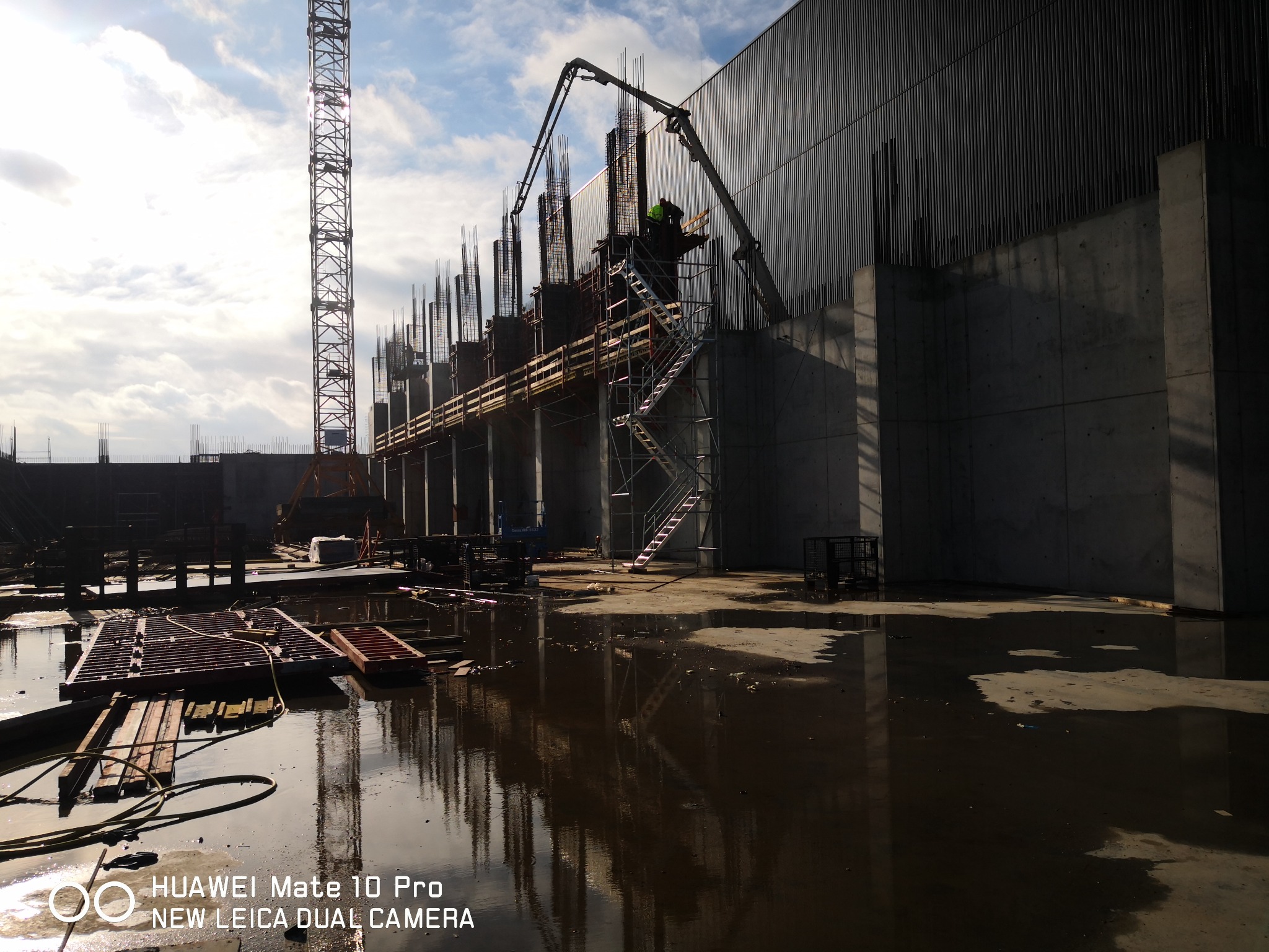 Budowa budynku żelbetowego w trakcie wylewania betonu, z widocznym dźwigiem i odbiciami w kałużach po deszczu.