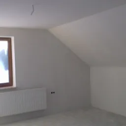 Poddasze po remoncie: białe ściany, okno z drewnianą ramą, grzejnik pod oknem, widoczne niedoskonałości w tynkowaniu.