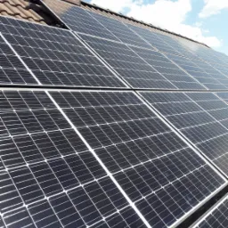 Panele słoneczne na dachu z ceglaną konstrukcją komina w tle, widoczne błękitne niebo z chmurami.