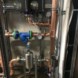 Kompleksowy system rur miedzianych z zaworami, manometrami i pompami Grundfos zamontowany na ścianie z bloków betonowych i szynach montażowych.