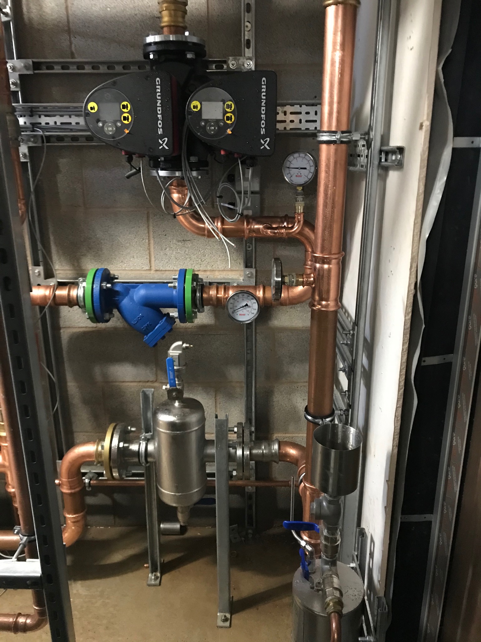 Kompleksowy system rur miedzianych z zaworami, manometrami i pompami Grundfos zamontowany na ścianie z bloków betonowych i szynach montażowych.