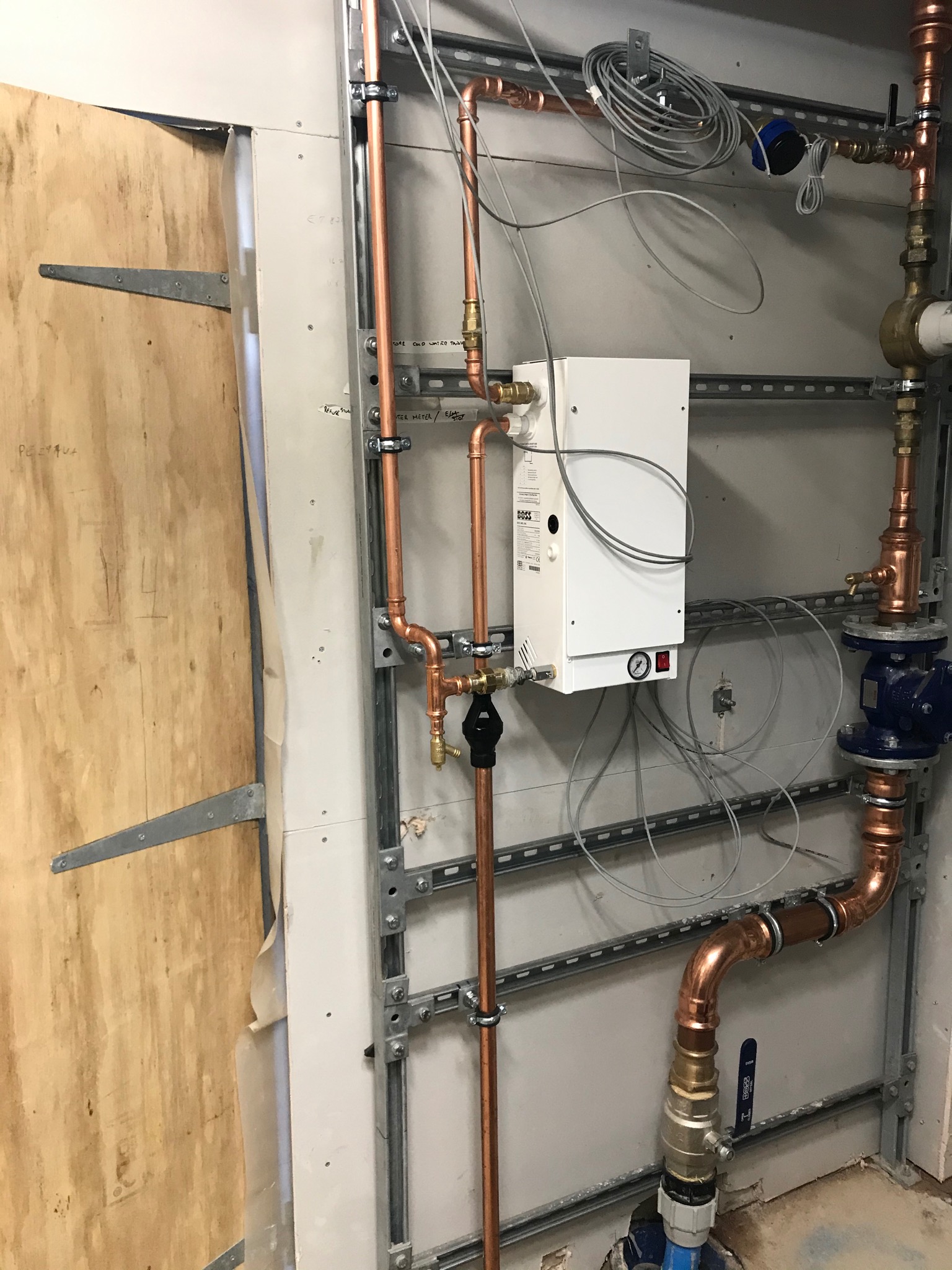 Nowa instalacja z miedzianych rur, zaworów i białego urządzenia grzewczego zamontowana na metalowej konstrukcji przy ścianie z płyt gipsowo-kartonowych. Widoczne liczne przewody i elementy mocujące.